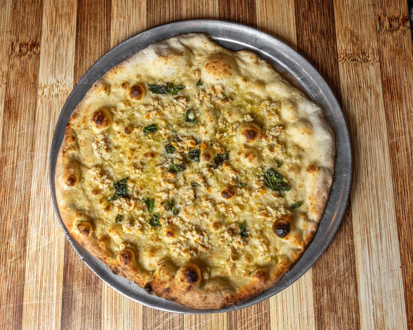 A white pizza with chicken, basil, and cheese on a metal pan, on a wooden table.