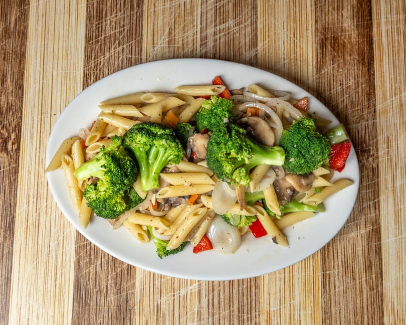 Penne pasta with broccoli, mushrooms, onions, and red bell peppers on a white oval plate.