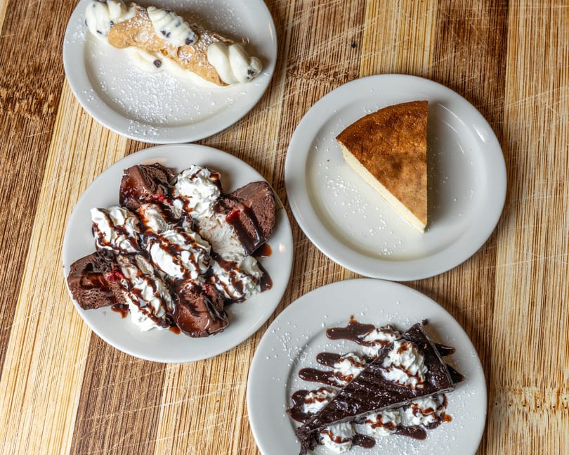Four dessert plates on a wooden table, including cannoli, cheesecake, and chocolate cake.