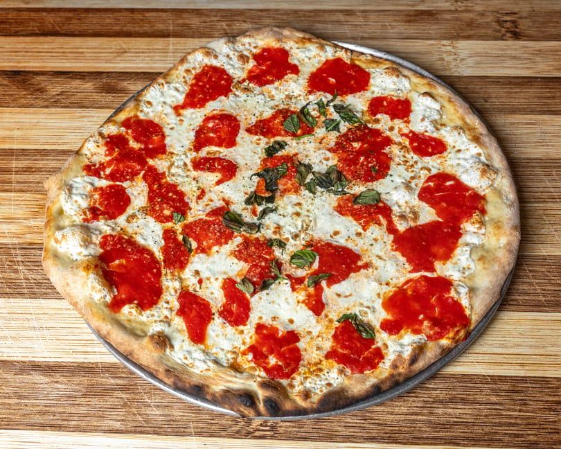 A freshly baked Margherita pizza with dollops of red sauce, white cheese, and basil on a wooden surface.