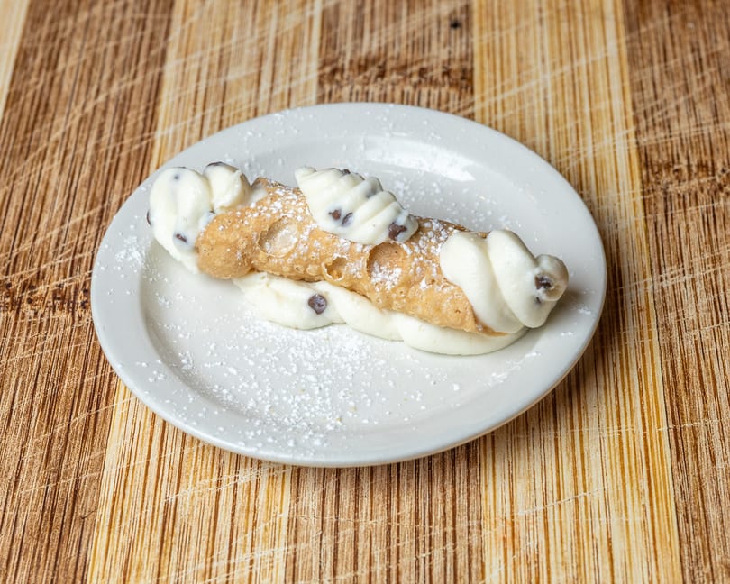 A cannoli with white cream filling and chocolate chips, dusted with powdered sugar on a white plate.