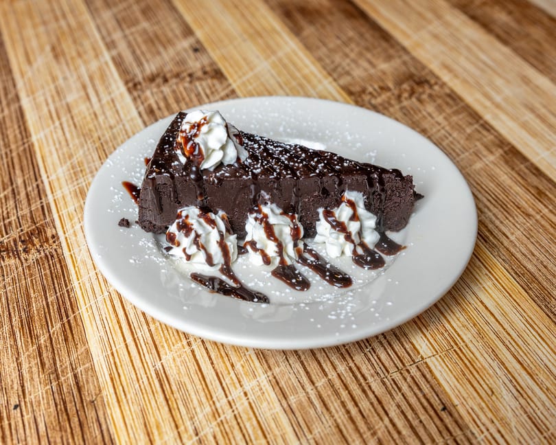A slice of rich chocolate cake with whipped cream, chocolate drizzle, and powdered sugar on a white plate.