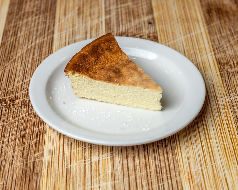 A slice of cheesecake with a golden-brown top and powdered sugar on a white plate.