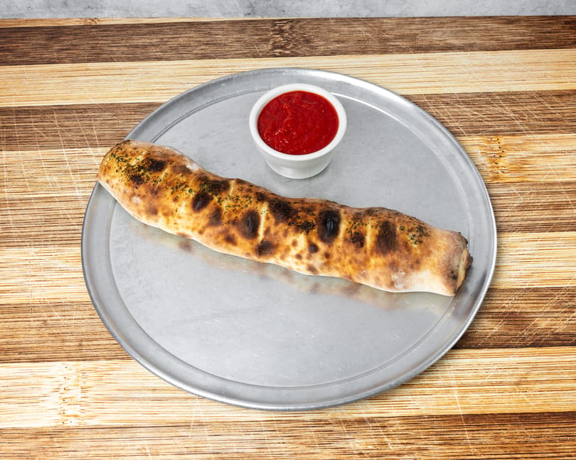 A golden-brown calzone with herbs on a metal pizza pan, served with a side of red marinara sauce.